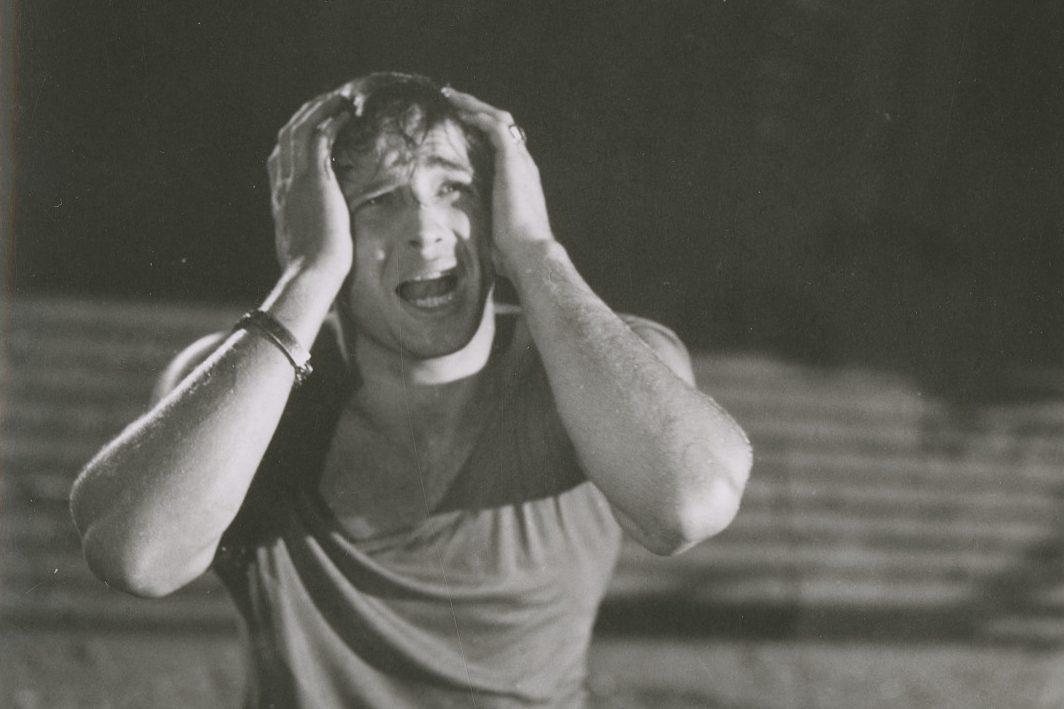 A person holds their head with both hands, expressing distress or frustration, in a black and white image. The background is blurred and dark, highlighting the individuals emotional expression.