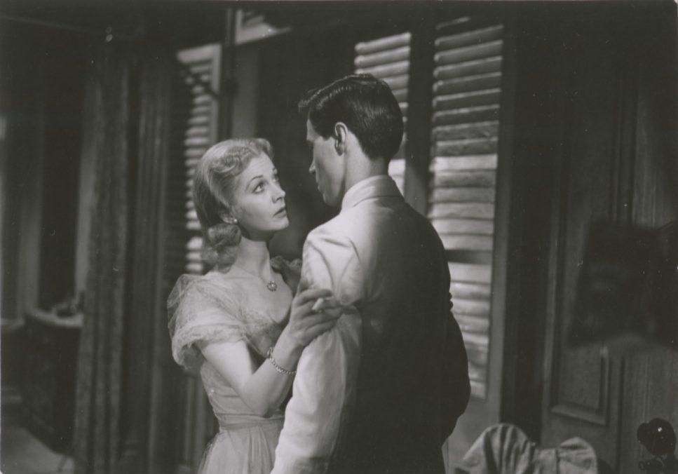 A woman in a vintage dress holds onto a mans arm in an intimate setting. They gaze at each other, standing near wooden shutters, creating a classic, dramatic atmosphere. The scene feels emotional and intense.