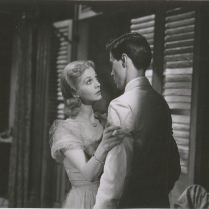 A black-and-white scene of a woman in a vintage dress, looking tenderly at a man in a suit. She gently holds his arm as they stand close together in front of partially open shutters.