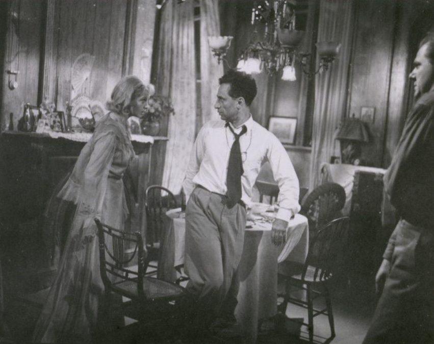 A vintage black and white scene in a dimly lit room shows two men and a woman having a tense conversation. The woman is standing on the left, while the two men face her. A table is set formally in the background.