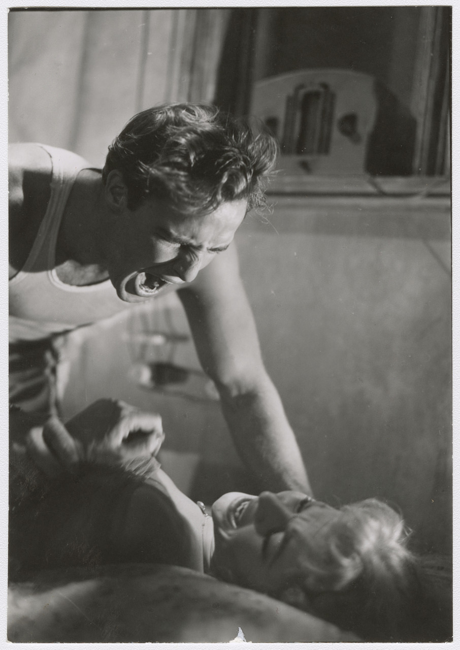 A black and white photo of a man leaning over a woman in a dramatic scene. The man appears to be yelling, while the woman looks up at him. The setting seems to be a dimly lit room with a radio in the background.