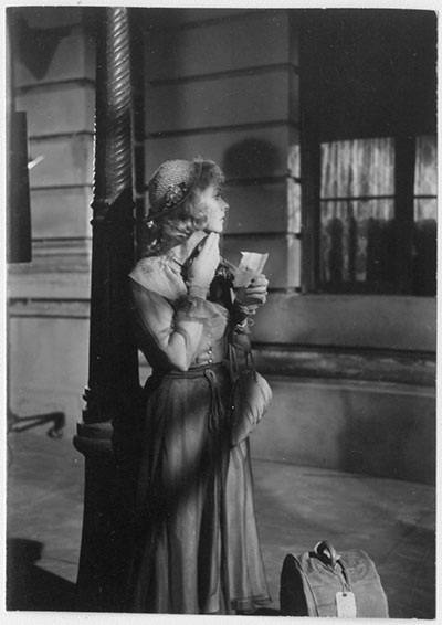A woman dressed in vintage attire stands in a dimly lit room, adjusting her makeup or hair while holding a compact mirror. She wears a hat and long gloves, with a small handbag and another bag on the floor nearby.