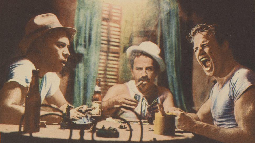 Three men sit around a table playing cards. Two wear hats, and one is smoking a cigar. The man on the right is shouting. The scene is dimly lit with drinks on the table.