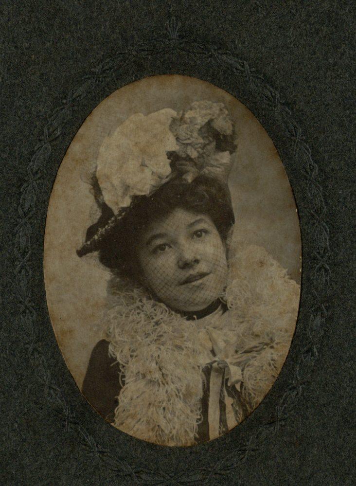 A vintage portrait of a woman wearing an ornate hat with large floral decorations. She has a fluffy scarf around her neck and is dressed in period attire, gazing directly at the camera. The photograph is enclosed in an oval frame.