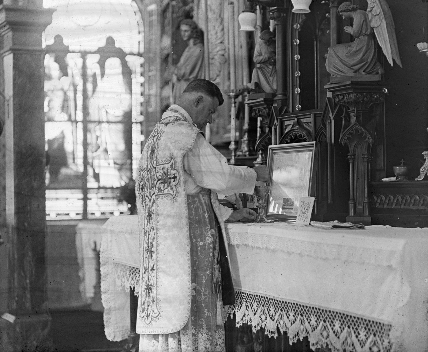 A black and white photo shows a Catholic priest at a church altar conducting Mass.