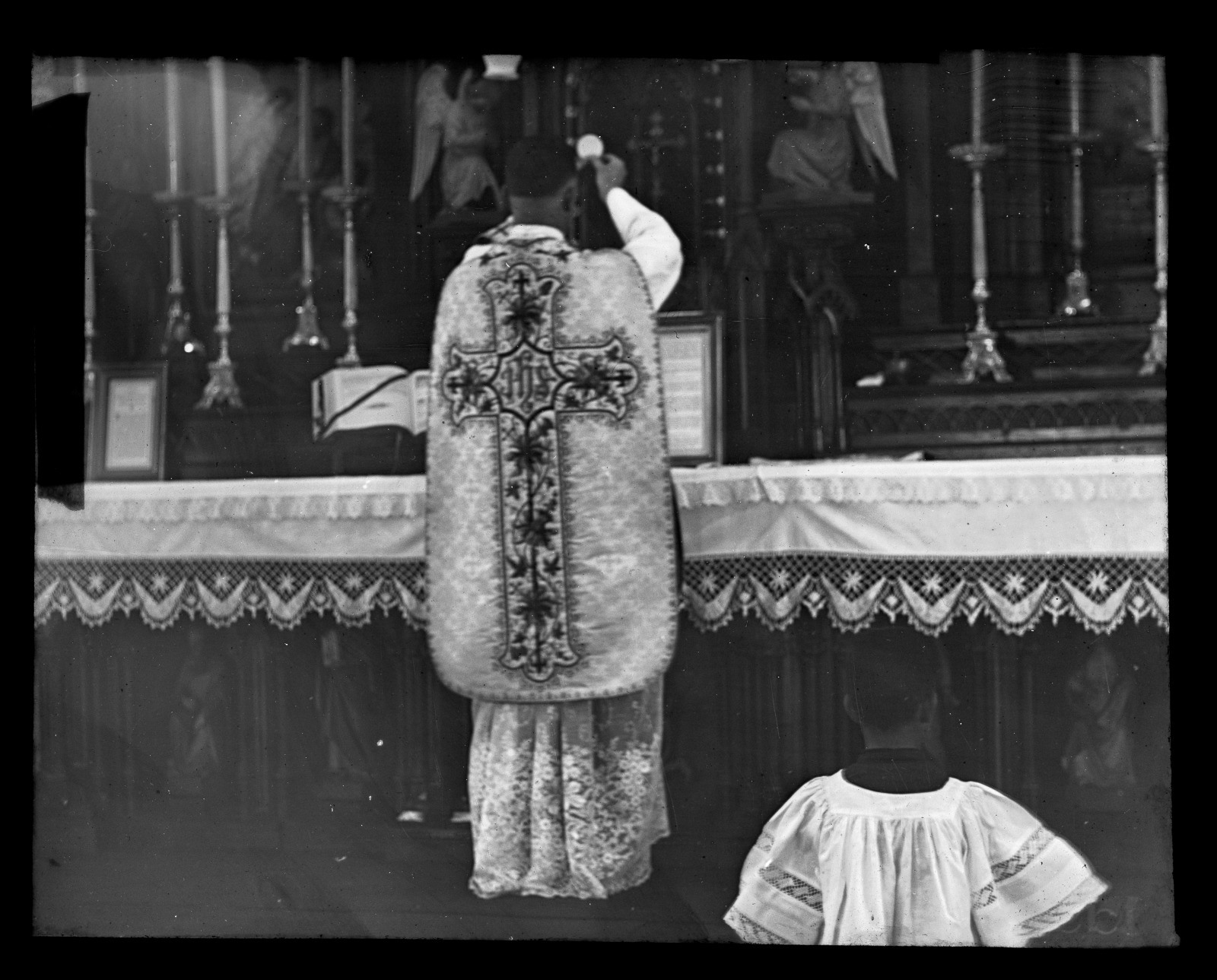 A priest performs a traditional religious ceremony at an ornate altar. He wears a decorated chasuble with a large cross on the back. An altar server kneels in the foreground, dressed in a white robe with dark accents.