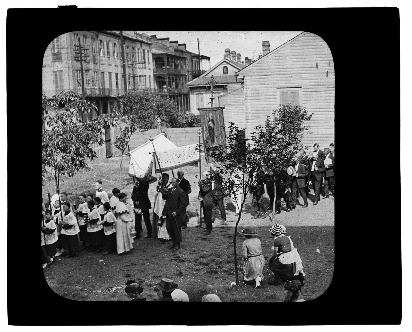 A black and white image showing a religious procession with people in robes and carrying banners. They walk through a neighborhood, passing wooden houses. Some participants hold candles, while onlookers gather on either side of the path.