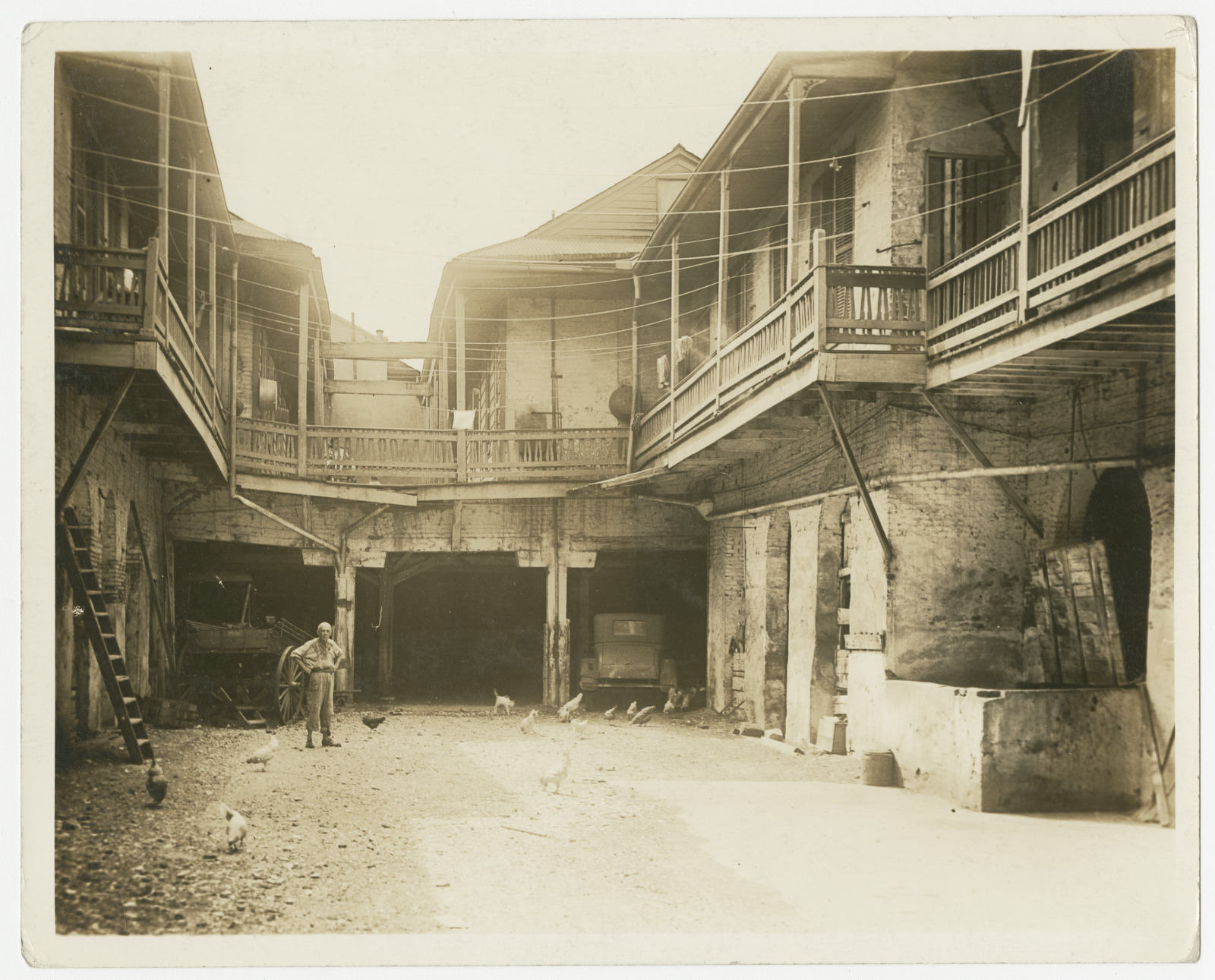 Black-and-white photograph of a courtyard at 919–925 Chartres Street, French Quarter, New Orleans during the 1930s. Shows a man standing with hands on hips in a large courtyard with wood galleries on the second story, clotheslines, a wagon, a car, chickens, cats, and a wooden ladder leaning against the building at left.