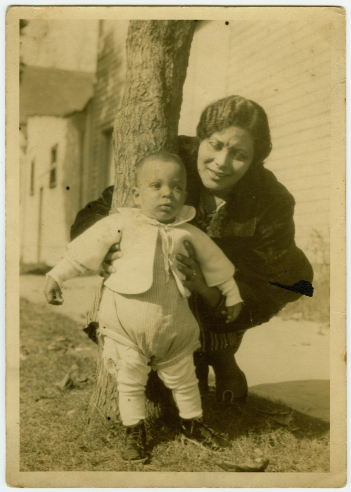 A vintage black and white photo of a woman crouching and smiling beside a toddler standing up against a tree. The toddler is wearing a white sweater and dark boots. A building and grass are visible in the background.
