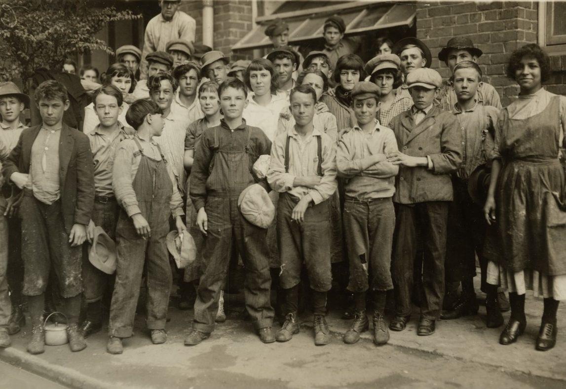 A black and white photo of a group of children, mostly boys, wearing early 20th-century work clothes. They stand closely together in front of a brick building, some holding hats. The children appear to be of various ages.