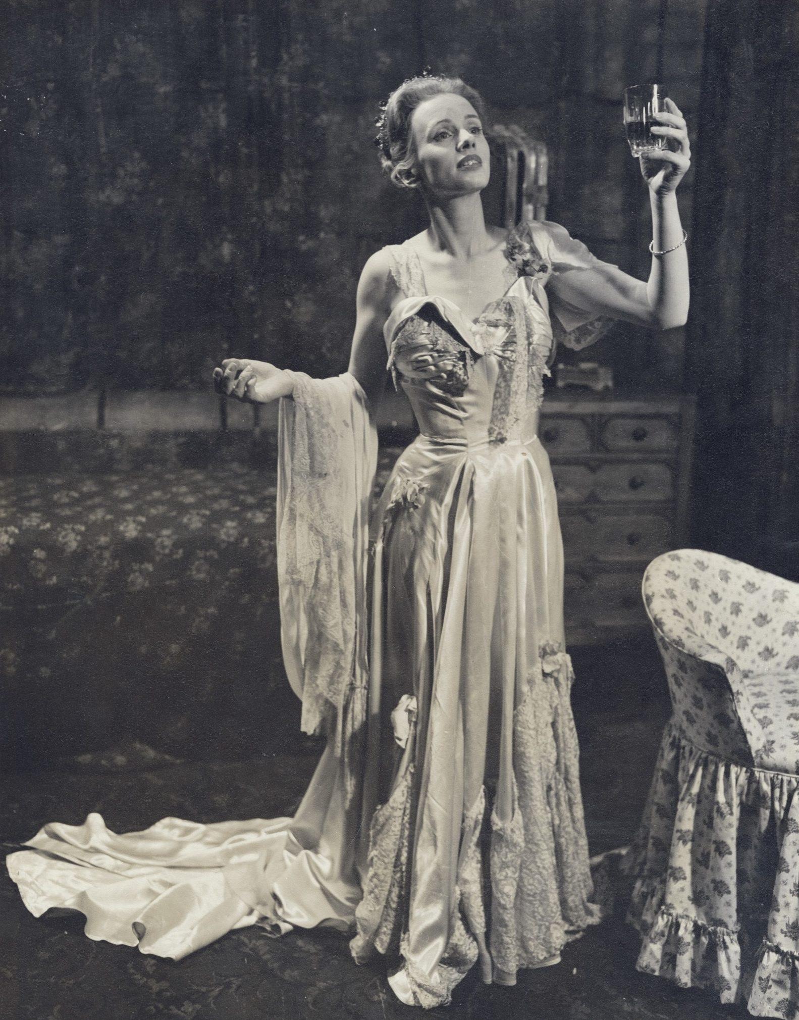 A woman in an elegant satin gown stands dramatically, holding a glass in one hand. Her other hand grips a lace shawl. The setting is a dimly lit room with a floral armchair and a small dresser in the background.