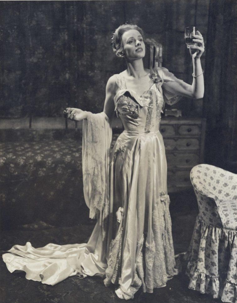 A woman in an elegant, flowing gown stands holding a glass aloft, her other arm outstretched. She is poised beside a plush armchair, with an ornate dresser in the background, exuding a theatrical or dramatic presence.