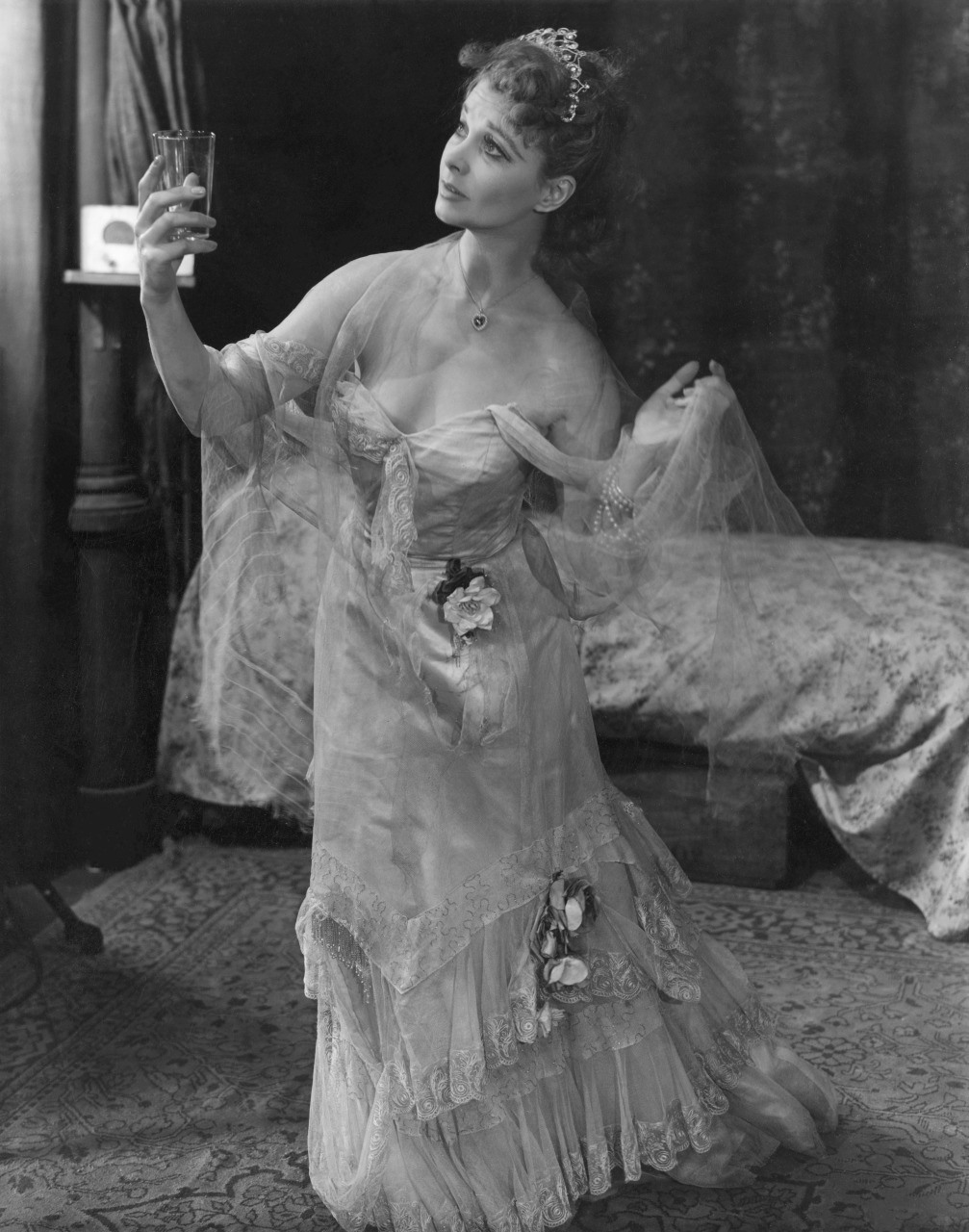 A woman in an ornate lace dress holds a glass aloft, gazing thoughtfully at it. She stands in a vintage setting with a bed in the background. Her dress is adorned with flowers, and she wears a small tiara. The scene is in black and white.
