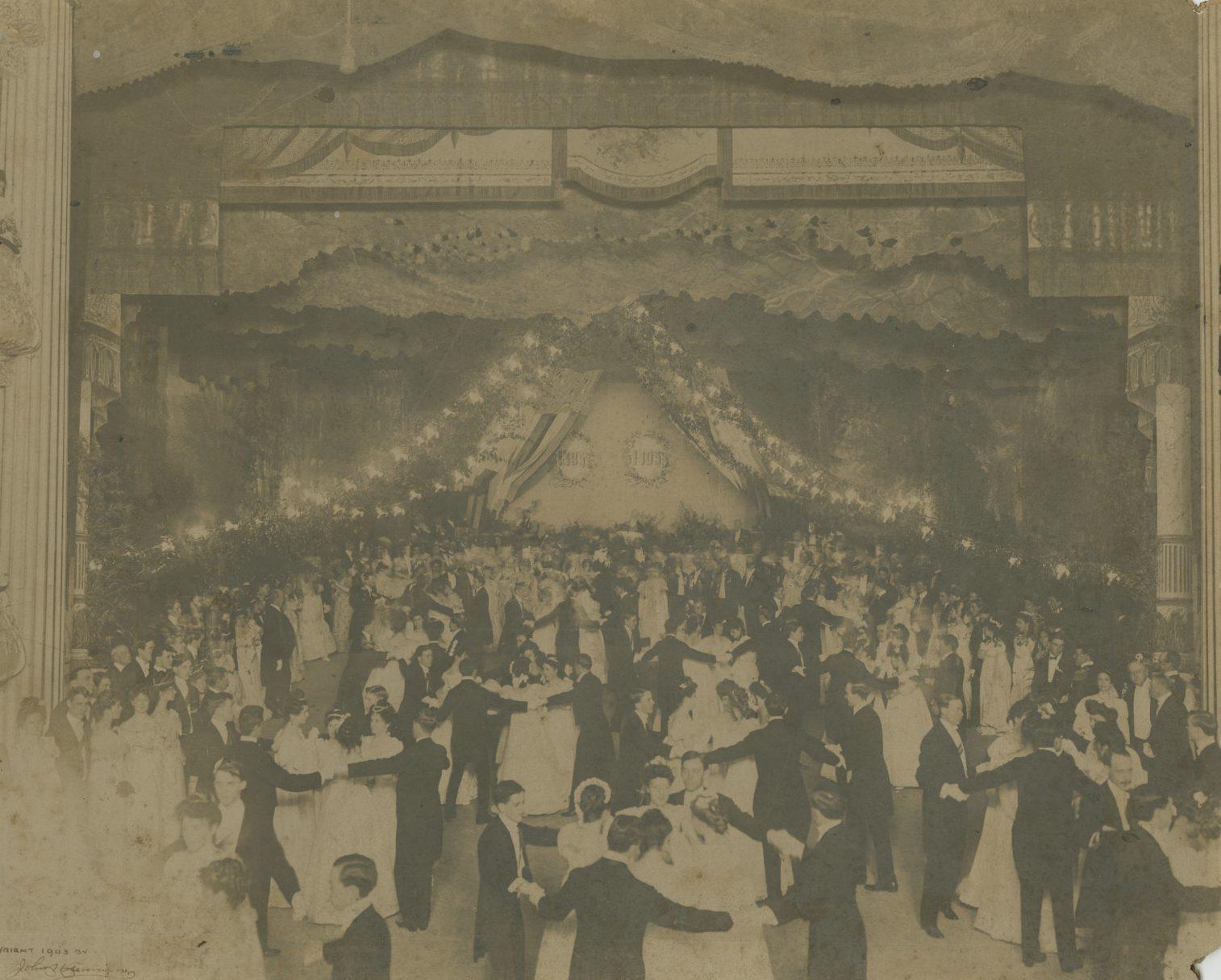 A vintage photograph of a grand ballroom dance. Numerous couples dressed in early 20th-century formal attire, with men in tuxedos and women in gowns, dance under elaborate decorations featuring garlands and drapery. The atmosphere is festive and elegant.