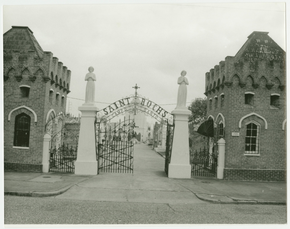 A black-and-white photo of the entrance to Saint Roch Cemetery. The image features an ornate wrought iron gate with the words Saint Roch and two brick towers with statues on top flanking the gate. The cemetery path leads into the distance.