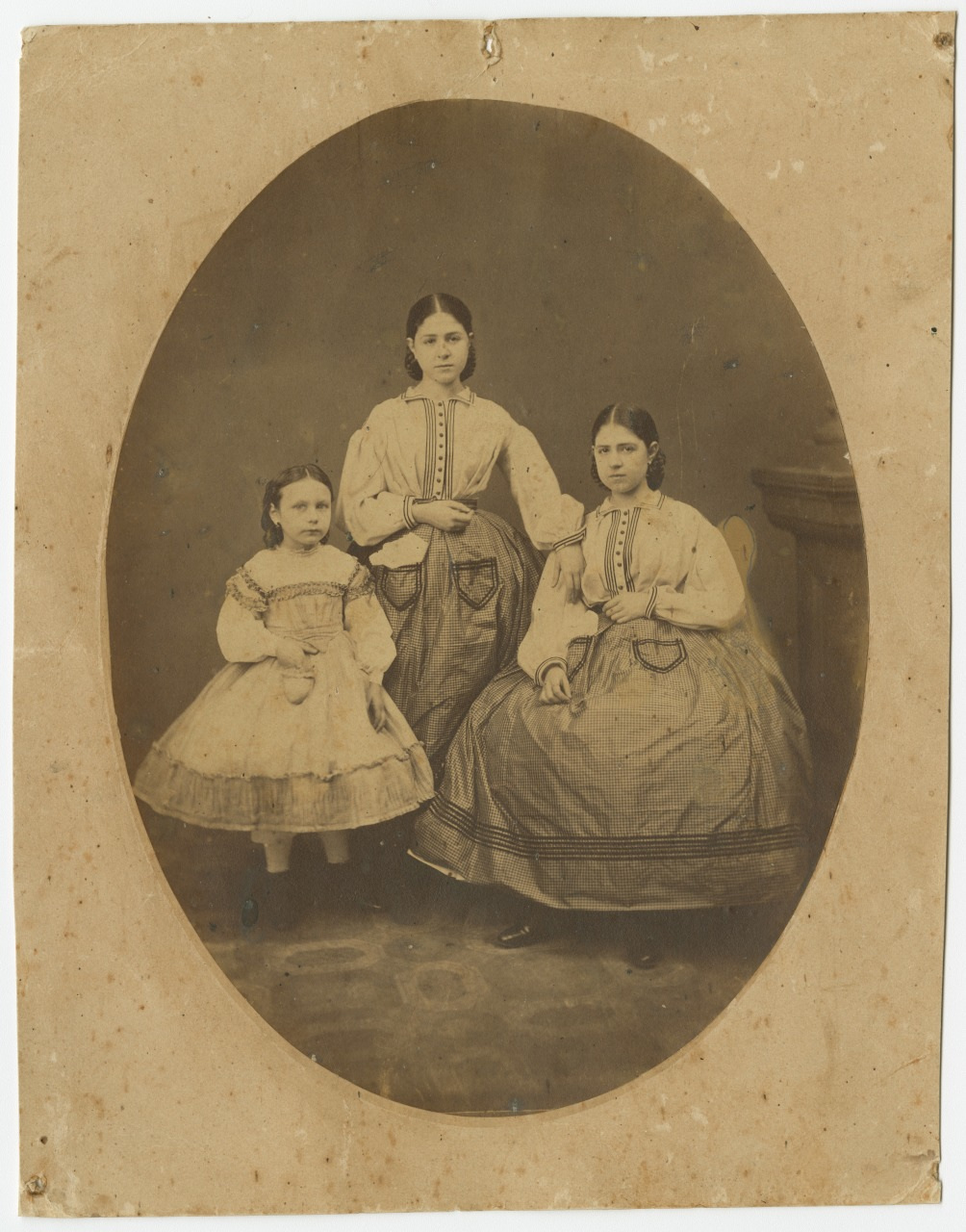 A sepia-toned vintage photograph of three young girls dressed in 19th-century attire. Two are standing while one is seated. They wear long-sleeved dresses with lace and ruffles, and their hair is styled neatly. The backdrop is simple and neutral.