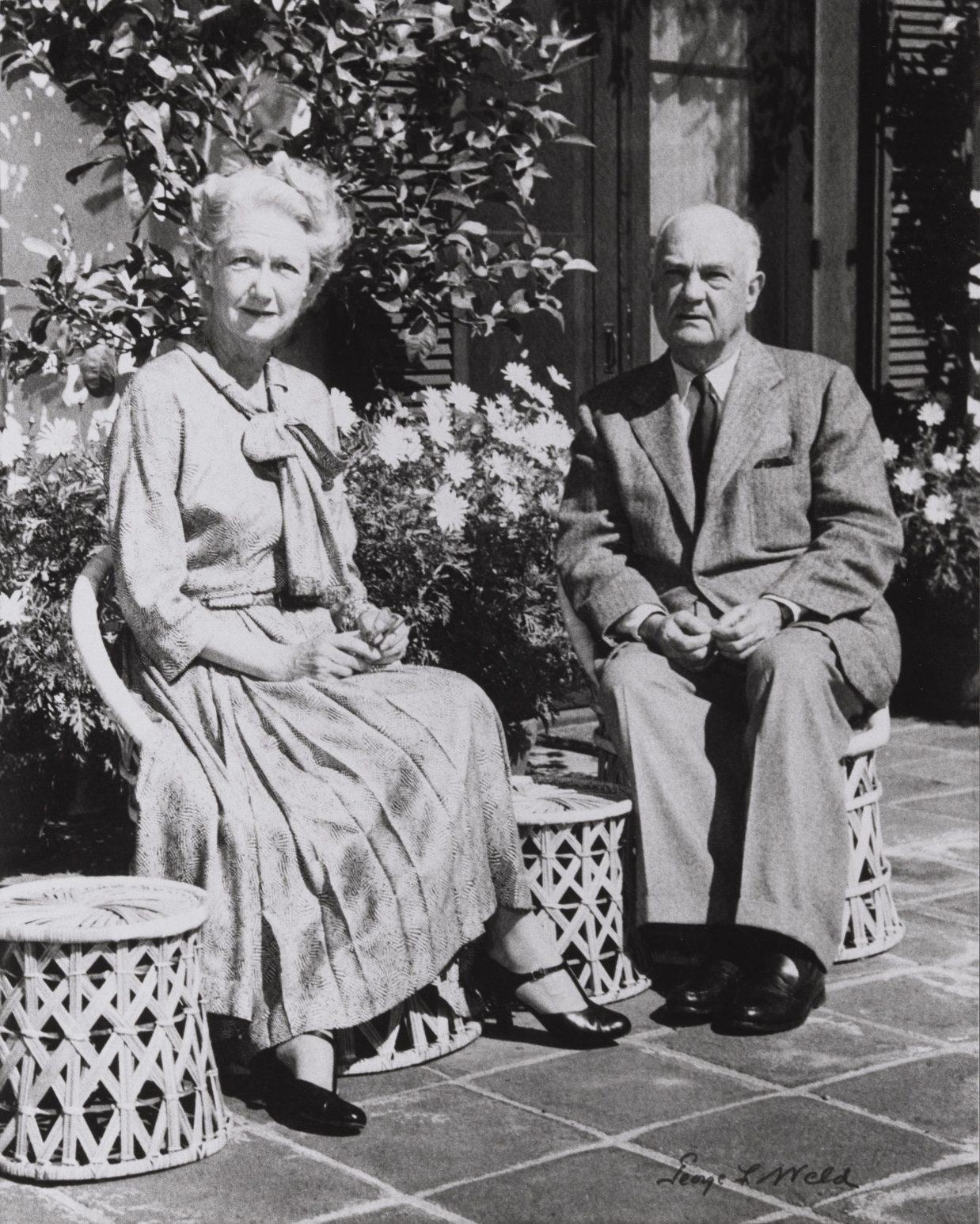 An elderly man and woman sit side by side on wicker stools outdoors. The woman wears a long dress with a bow, and the man is in a suit. They are surrounded by plants and appear relaxed.
