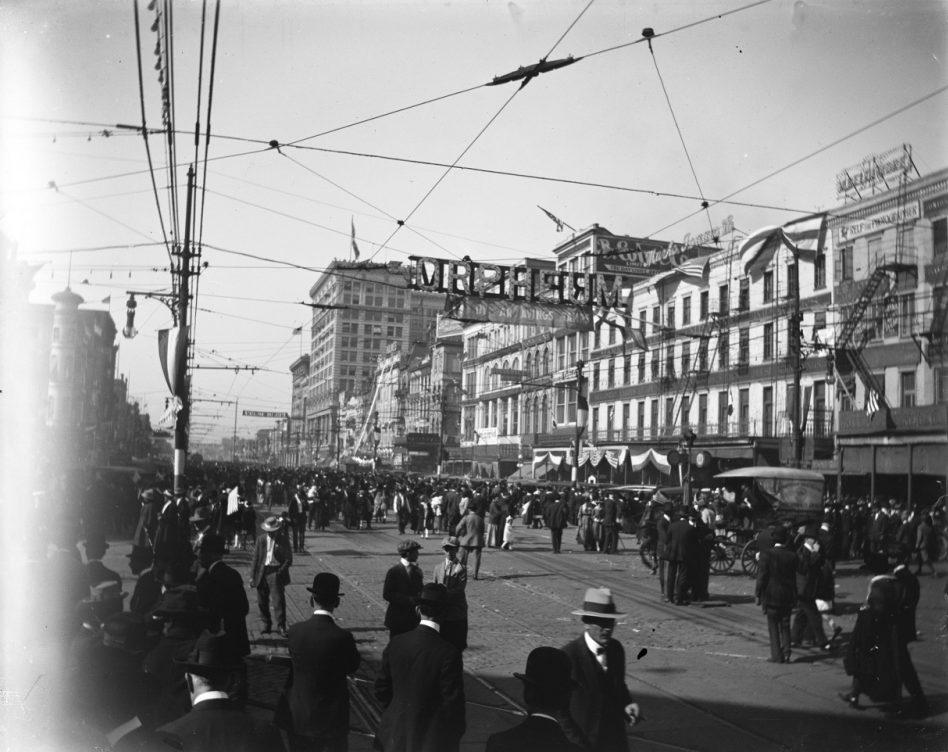 A bustling city street scene from the early 1900s, featuring crowds of people in period attire. Streetcar tracks run through the road, flanked by multi-story buildings adorned with signs and banners. Overhead, wires crisscross and a banner reads Mr. Erect.