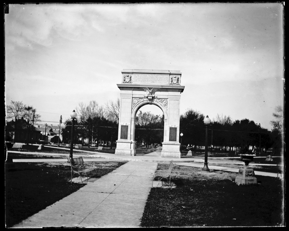 Black and white image of a large stone archway in a park setting. Pathways lead up to it, flanked by benches and lamp posts. Trees are visible in the background under a cloudy sky.