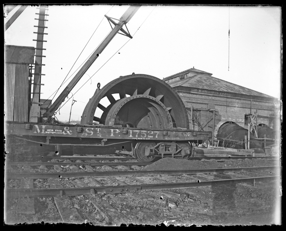 A vintage black and white photograph showing a large industrial wheel on a flatbed train car with M. & St. P. 17627 written on its side. The train is on rail tracks, with a building and another wheel in the background.