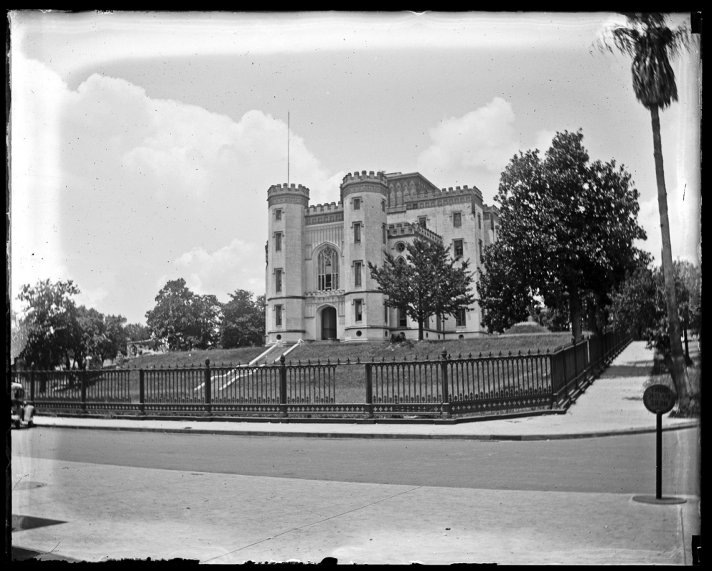 A historic stone castle-like building with turrets and arched windows, surrounded by a wrought iron fence. The building is set on a grassy hill with trees, under a partly cloudy sky. A street and a vintage car are visible in the foreground.