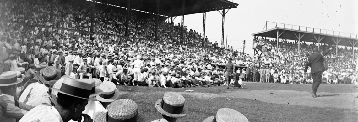 A vintage black-and-white photo of a large crowd at a baseball game. Spectators, mostly in hats, fill the stands, with some people seated on the ground. A player stands ready to pitch on the field. The stadium structure is visible.