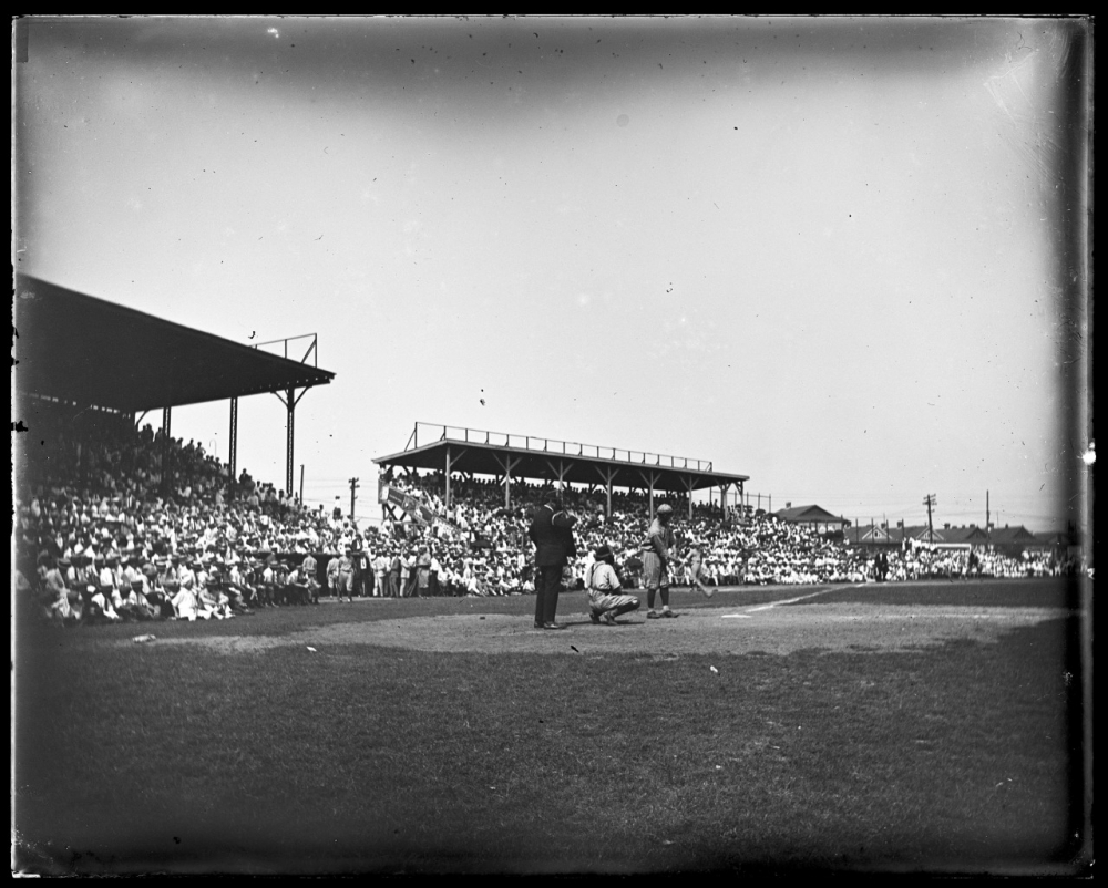 A vintage baseball game scene with a batter at the plate, a catcher, and an umpire. The stands are packed with spectators on a sunny day. The ballpark background includes grandstands and a clear sky.