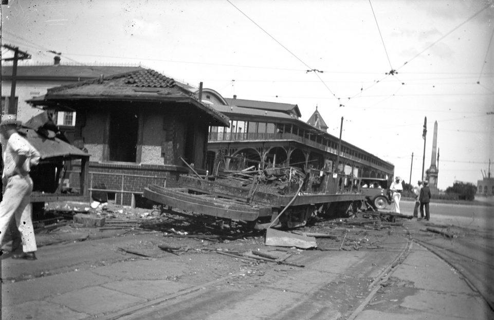 A damaged streetcar lies wrecked on the tracks, surrounded by debris. A small building nearby shows signs of damage. Several people stand around observing the scene. Overhead wires and power poles are visible against a cloudy sky.