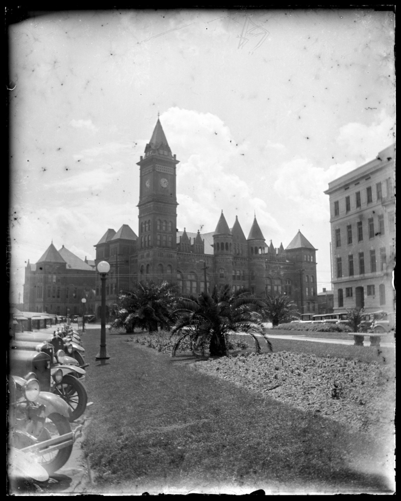 A black and white photograph of a historic building with a tall clock tower, featuring Gothic architecture. In the foreground, theres a manicured lawn with palm trees and vintage cars parked along the street. The sky is clear and bright.