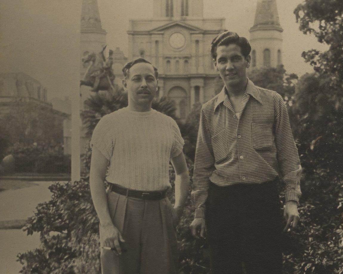 Two men stand in a garden with a historic building in the background, featuring tall towers and a clock. One man wears a light sweater and trousers, the other a checkered shirt and dark pants. Both hold cigars and appear relaxed.