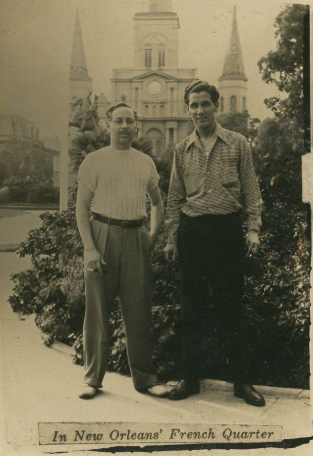 Two men stand in front of a historic building in the French Quarter, New Orleans. The building has tall spires and a large clock. One man wears a short-sleeve shirt and trousers, the other a long-sleeve shirt and trousers. The caption reads, In New Orleans French Quarter.
