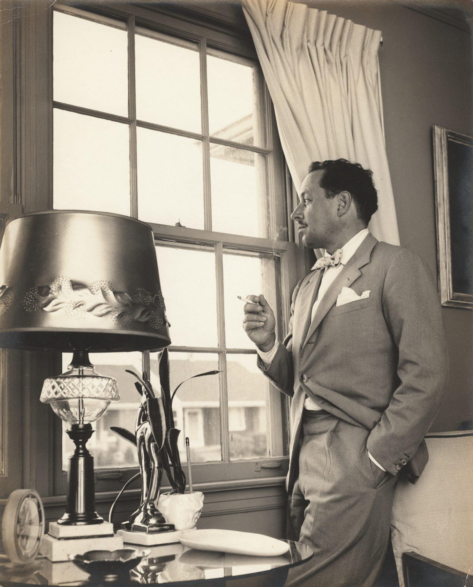 A man in a suit stands by a window, holding a cigarette. He appears thoughtful, looking outside. A lamp and a small decorative plant are on the table nearby. Curtains frame the window, and the room is softly lit.