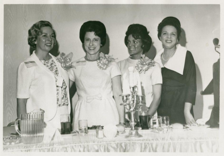 Four women stand behind a table set with drinks and a candle holder. They are dressed in 1960s fashion, with two wearing light dresses and corsages, and two in dark outfits. All are smiling, captured in black and white.