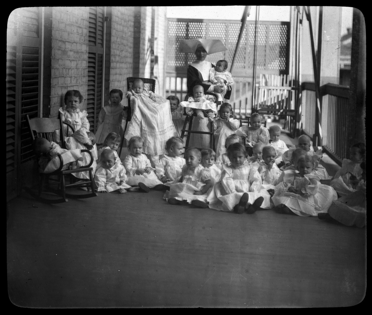 A group of toddlers and babies in white outfits sit and stand on a porch. Two women, one holding a baby, oversee the children. The porch has a wooden floor, brick wall, and slatted windows. Some children are seated in wooden chairs.