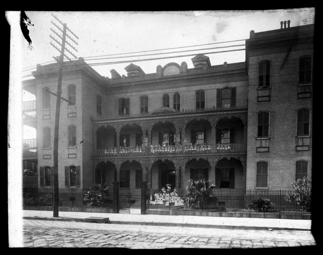 A historic brick building with ornate balconies and detailed ironwork features prominently in this black and white photo. It is flanked by palm trees and sits behind a wrought iron fence, with cobblestone street and utility poles in the foreground.