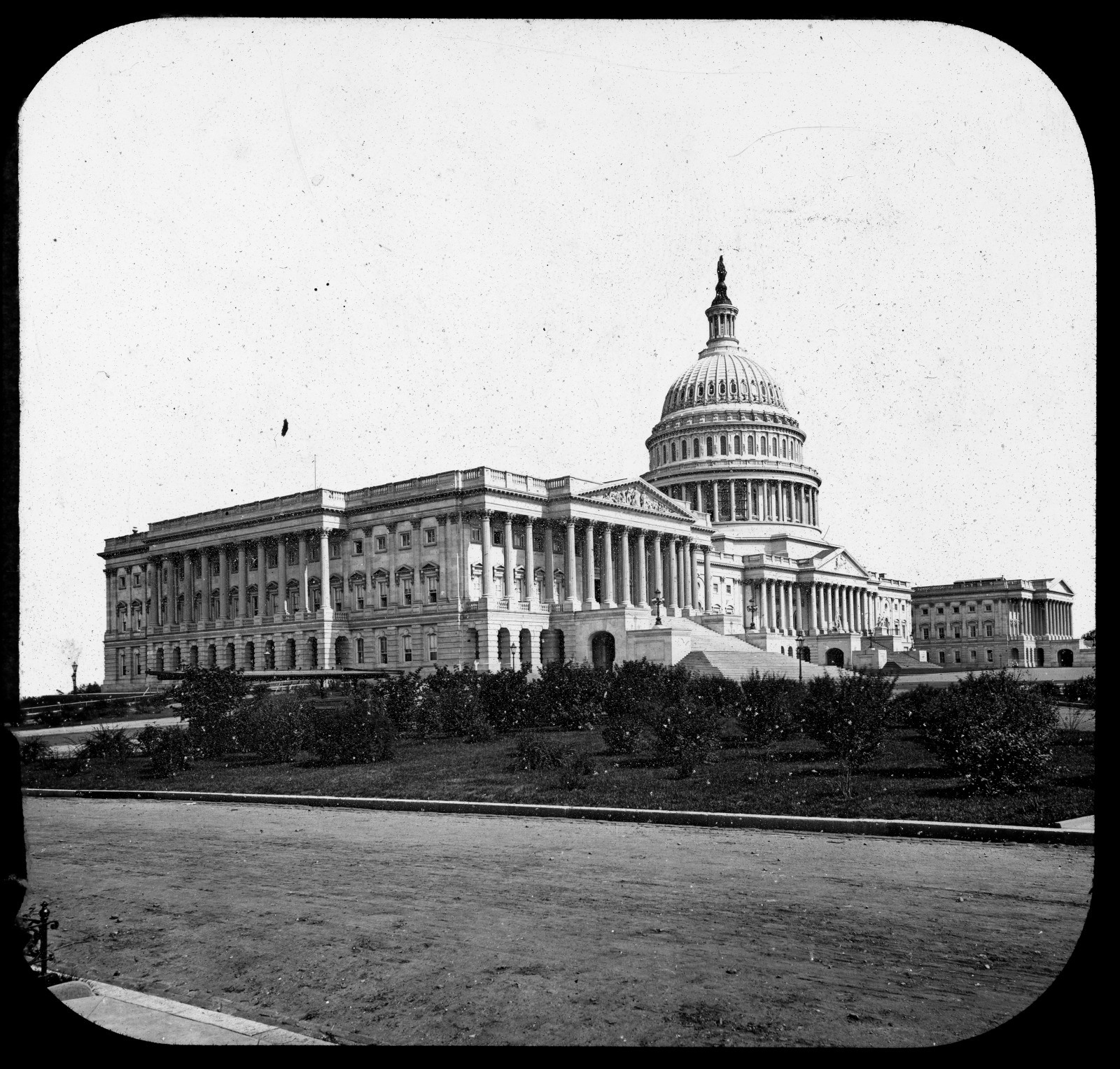 A vintage black-and-white photograph of the United States Capitol building, featuring its iconic dome and neoclassical architecture. The foreground shows a path and some trees. The sky is clear.