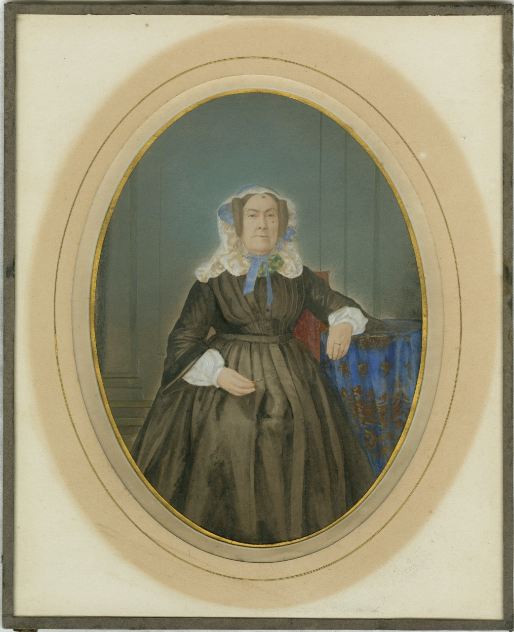 A Victorian-era portrait of an elderly woman seated, wearing a dark dress and a lace bonnet. She rests one arm on a table draped with an ornate blue cloth. The image is framed in an oval border.