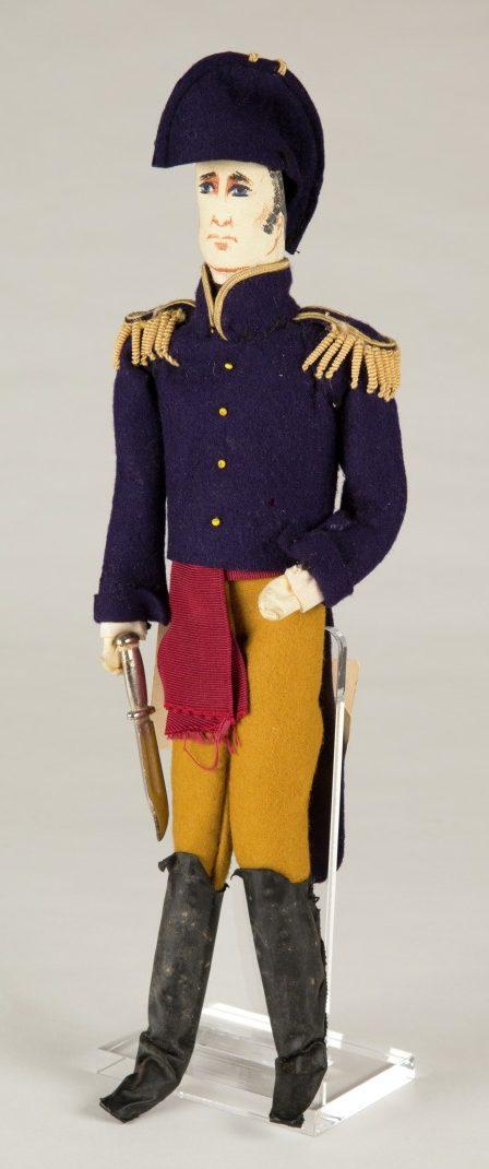 A fabric doll of a soldier wearing a dark purple military-style coat with gold shoulder epaulettes, red and mustard pants, and black boots. The doll holds a sword and wears a matching purple hat.