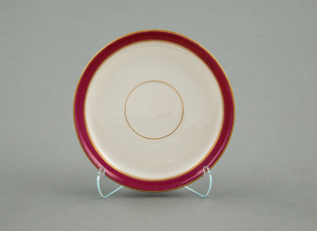 A decorative porcelain plate with a cream base and a wide, deep red border edged with gold. The plate is displayed on a clear stand against a gray background.
