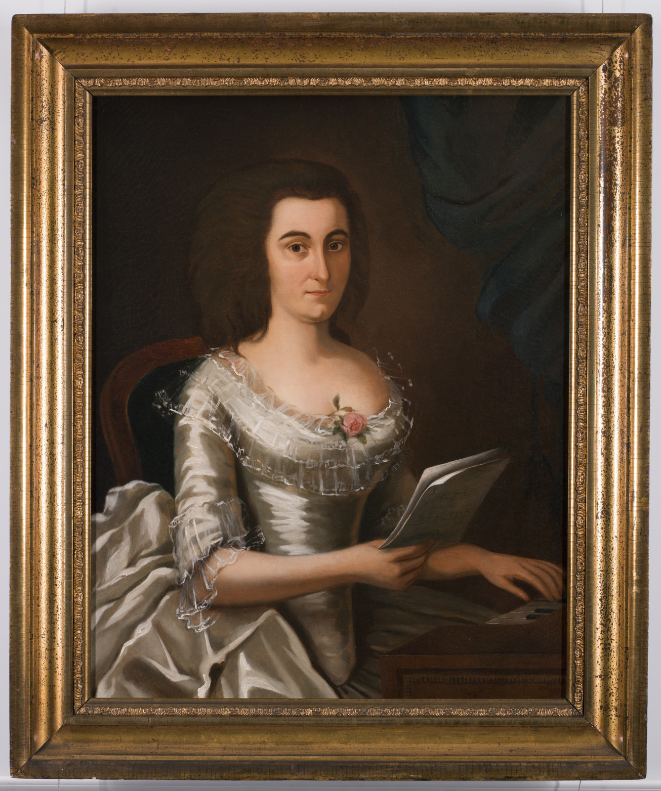 Painting of a woman in 18th-century attire seated at a table, holding a sheet of paper. She wears a white, lacy dress adorned with a pink rose. The background is a dark, muted shade. The portrait is framed in an ornate gold frame.