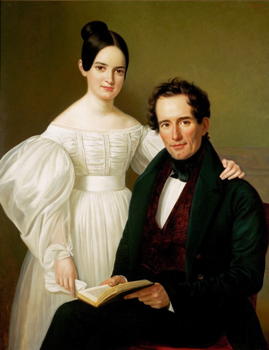 A 19th-century portrait of a seated man and a standing woman. The man, holding an open book, wears an elegant dark suit. The woman, in a white dress with large puffed sleeves, rests her hand on the mans shoulder. Both are looking forward.