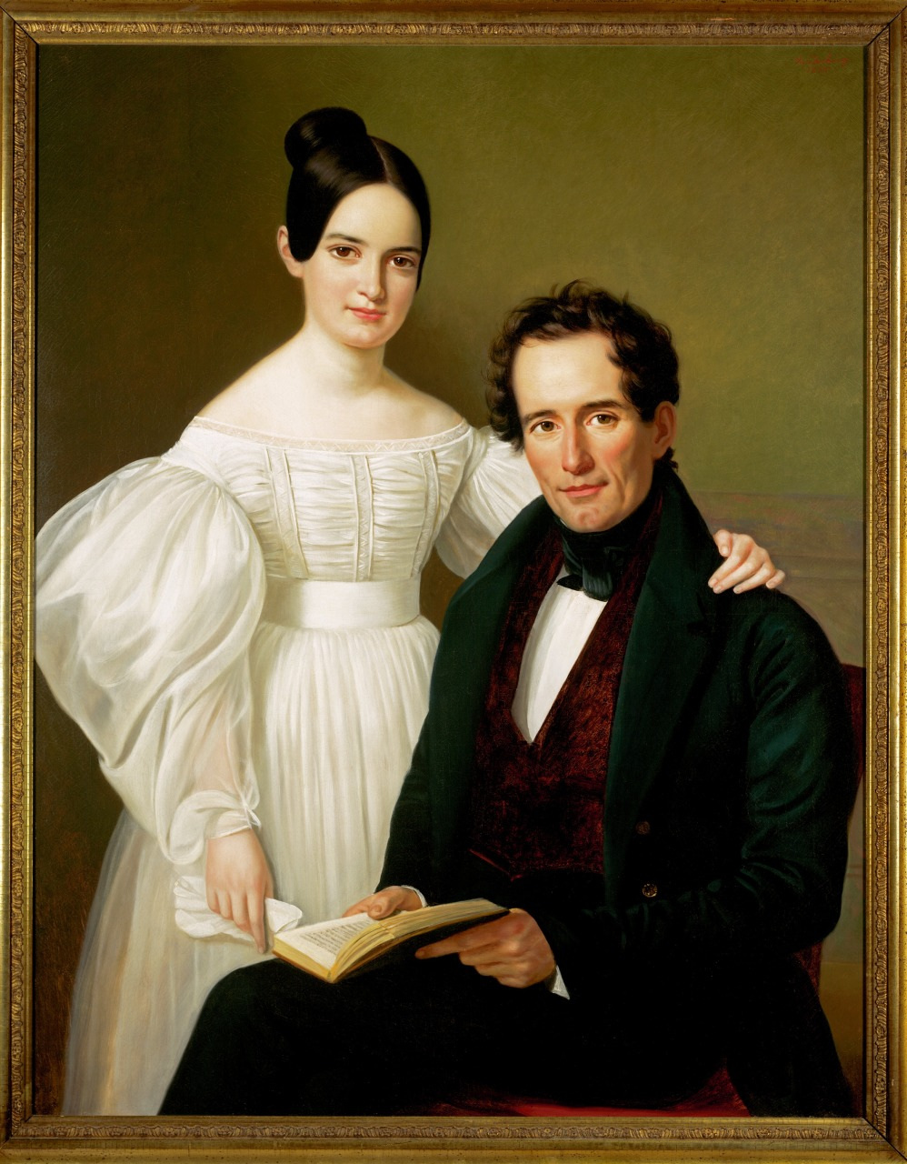 A 19th-century portrait of a couple. A woman in a white dress stands with her arm on the shoulder of a seated man in a dark suit holding a book. They face the viewer against a neutral background. Framed in an ornate gold frame.