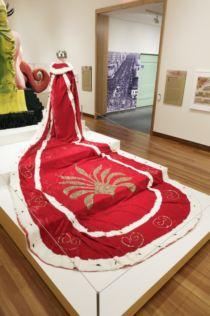 A regal red and white velvet robe with a crown displayed in a museum. The robe features gold embroidery and a long, flowing train, positioned on a platform with various artworks in the background.