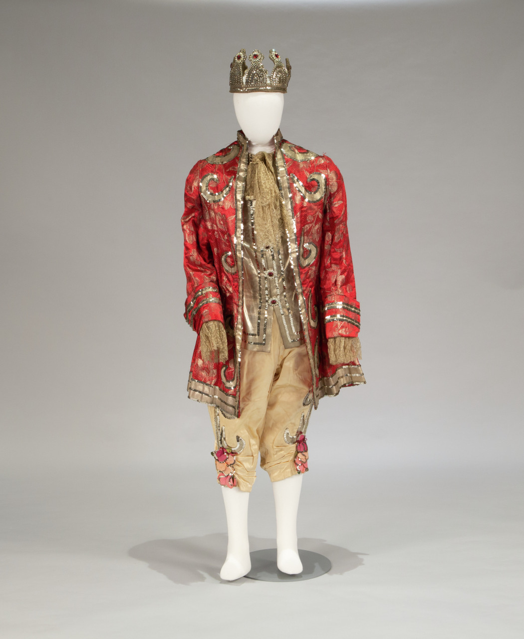 A mannequin displays an elaborate costume featuring a gold crown, a red embroidered coat with intricate designs, a gold scarf, and matching knee-length breeches adorned with floral details. The outfit exudes a regal and theatrical style.