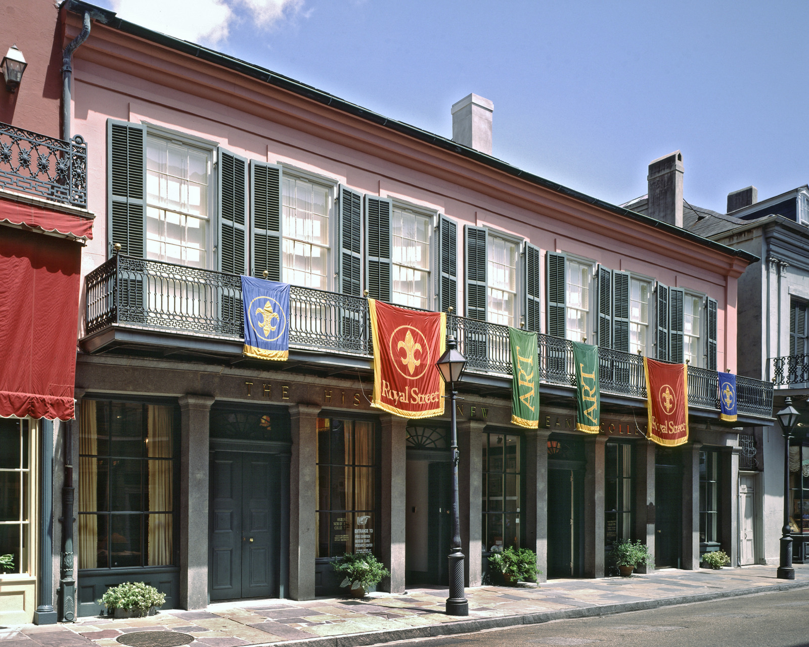A historic two-story building with pink walls and green shutters, adorned with red and blue banners. The architectural style is classic, with arched windows and a balcony. The street is cobblestone, adding to the vintage atmosphere.