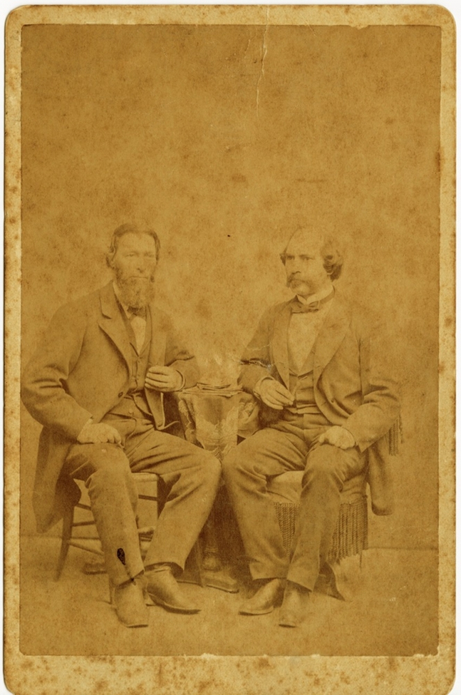 A sepia-toned vintage photograph depicts two bearded men in 19th-century suits seated beside a small table. The background and details show signs of aging and wear, adding to the antique aesthetic of the image.