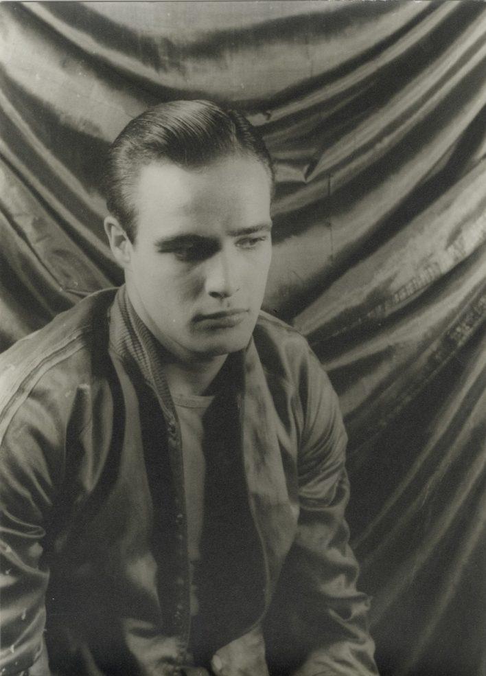 A black and white portrait of a man with slicked-back hair, wearing a jacket over a shirt. He appears deep in thought, seated against a softly draped fabric background.