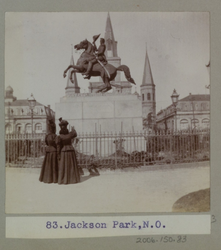 A vintage photograph depicting two women in long dresses standing in front of an equestrian statue on a raised pedestal, surrounded by an ornate fence. In the background, there are two pointed church spires and historic buildings.