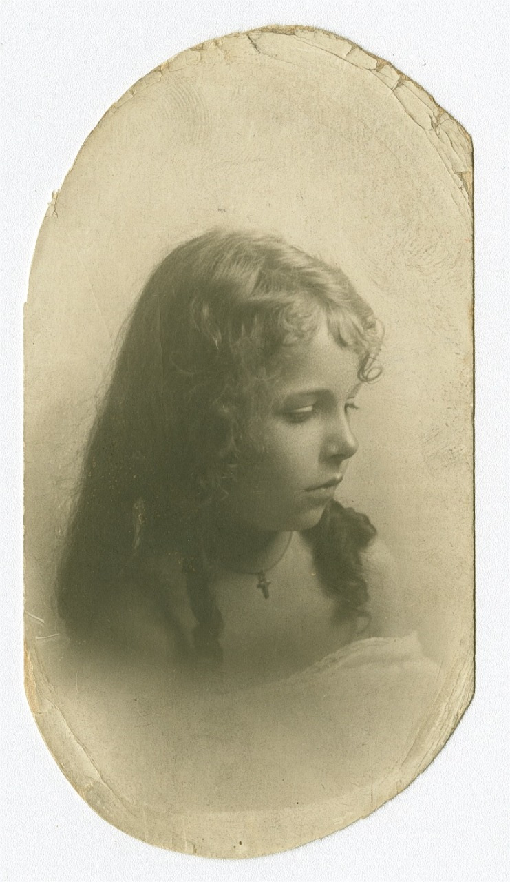 Sepia-toned vintage portrait of a young girl with curly hair, looking down with a gentle expression. She wears a dark necklace with a small cross pendant. The oval edges of the photograph are slightly worn.
