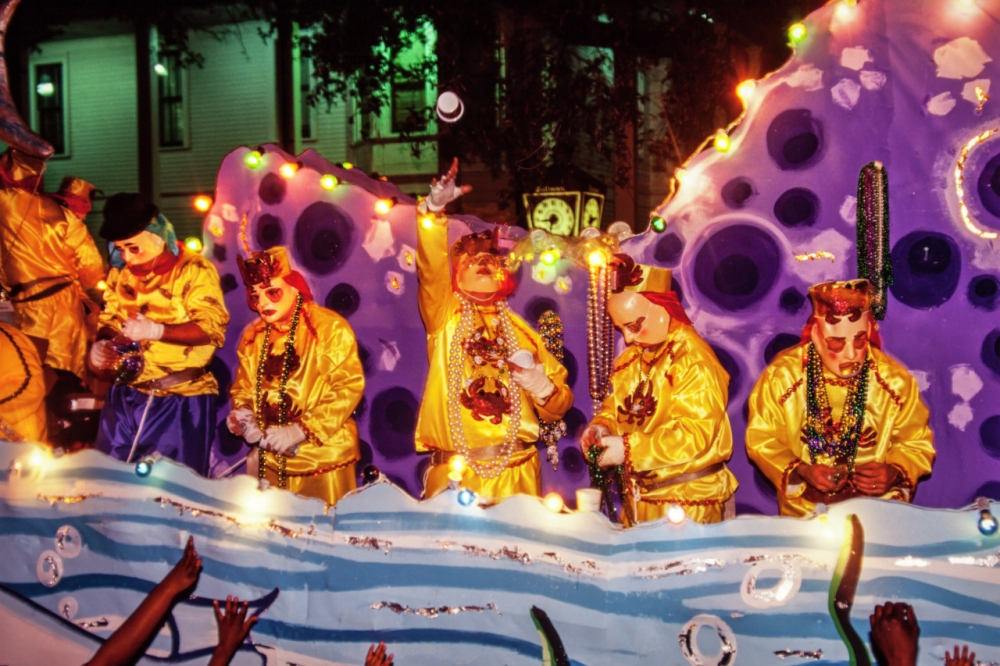 People in colorful costumes and masks stand on a brightly decorated float during a nighttime parade. They are wearing vibrant yellow outfits and throwing beads to the crowd. The float is adorned with lights and decorations.